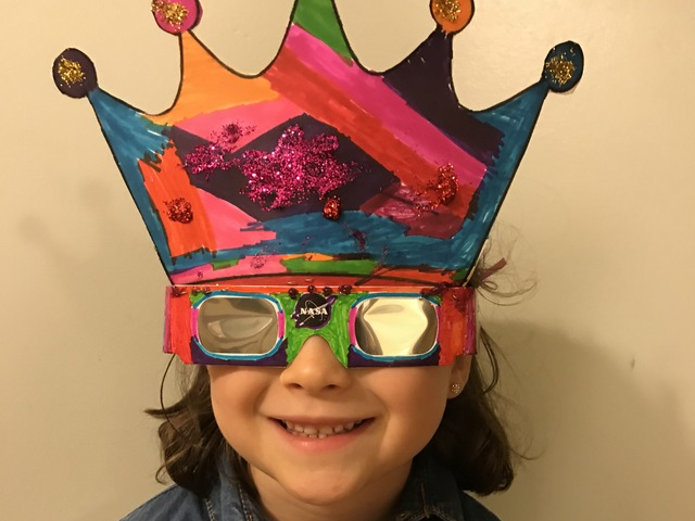 A child wears eclipse glasses that are colored in with markers and have a colorful crown with glitter attached.
